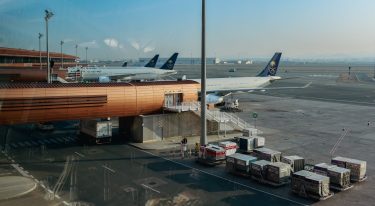 Repülőgépek állnak a kifutón, miközben rakodóegységek sorakoznak a repülőtér előterében.