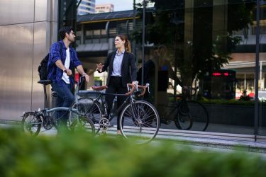 Két ember beszélget kerékpárral modern városi környezetben, ami a rövid elérésre épülő, fenntartható közlekedési szemléletet illusztrálja.