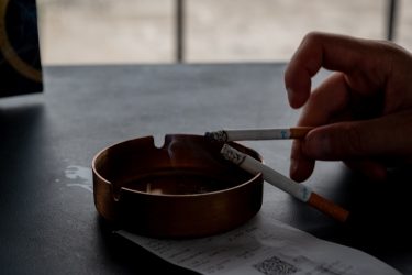 Kéz tart cigarettát egy hamutartó fölött, ami a kültéri dohányzóhelyek tisztasági problémáira utal.