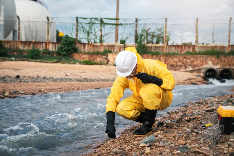 Védőruhás szakember vízmintát vesz egy ipari terület közelében húzódó vízfolyásból.