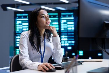 Irodai környezetben dolgozó nő monitor előtt, háttérben szerverekkel és digitális infrastruktúrával.