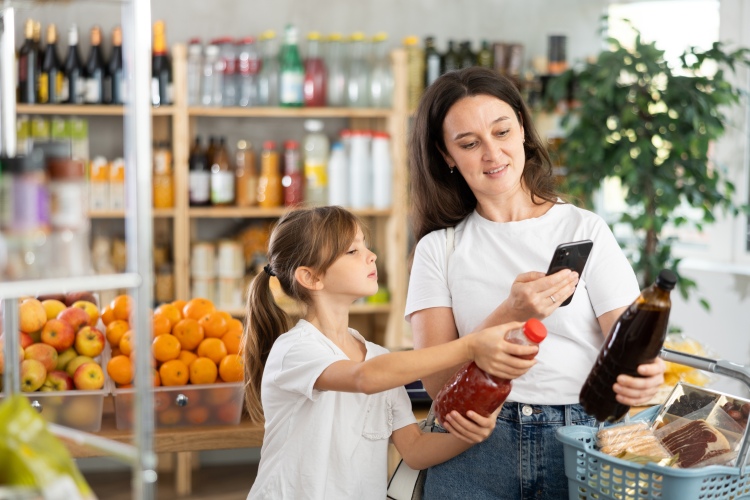 Egy nő és egy gyermek boltban terméket tartanak, miközben mobiltelefonnal információt keresnek a csomagoláson.